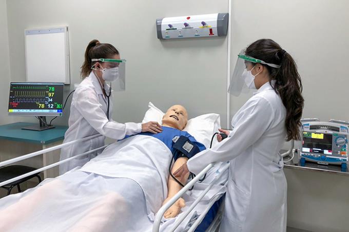 Duas estudantes de medicina, usando jalecos, máscaras e protetores faciais, examinam um manequim de paciente em uma cama de hospital. Uma estudante usa um estetoscópio enquanto a outra verifica a pressão arterial do manequim. Um monitor cardíaco e outros equipamentos médicos são visíveis ao fundo.