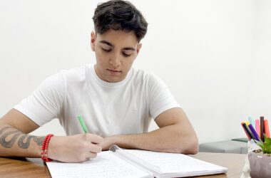 Jovem estudante sentado à mesa durante a preparação para o vestibular, escrevendo em um caderno com caneta verde. Ao lado, um suporte com canetas coloridas e uma pequena planta ajudam a compor um ambiente de estudo organizado e acolhedor, ideal para manter o foco e a disciplina.