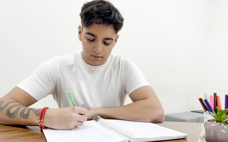 Jovem estudante sentado à mesa durante a preparação para o vestibular, escrevendo em um caderno com caneta verde. Ao lado, um suporte com canetas coloridas e uma pequena planta ajudam a compor um ambiente de estudo organizado e acolhedor, ideal para manter o foco e a disciplina.