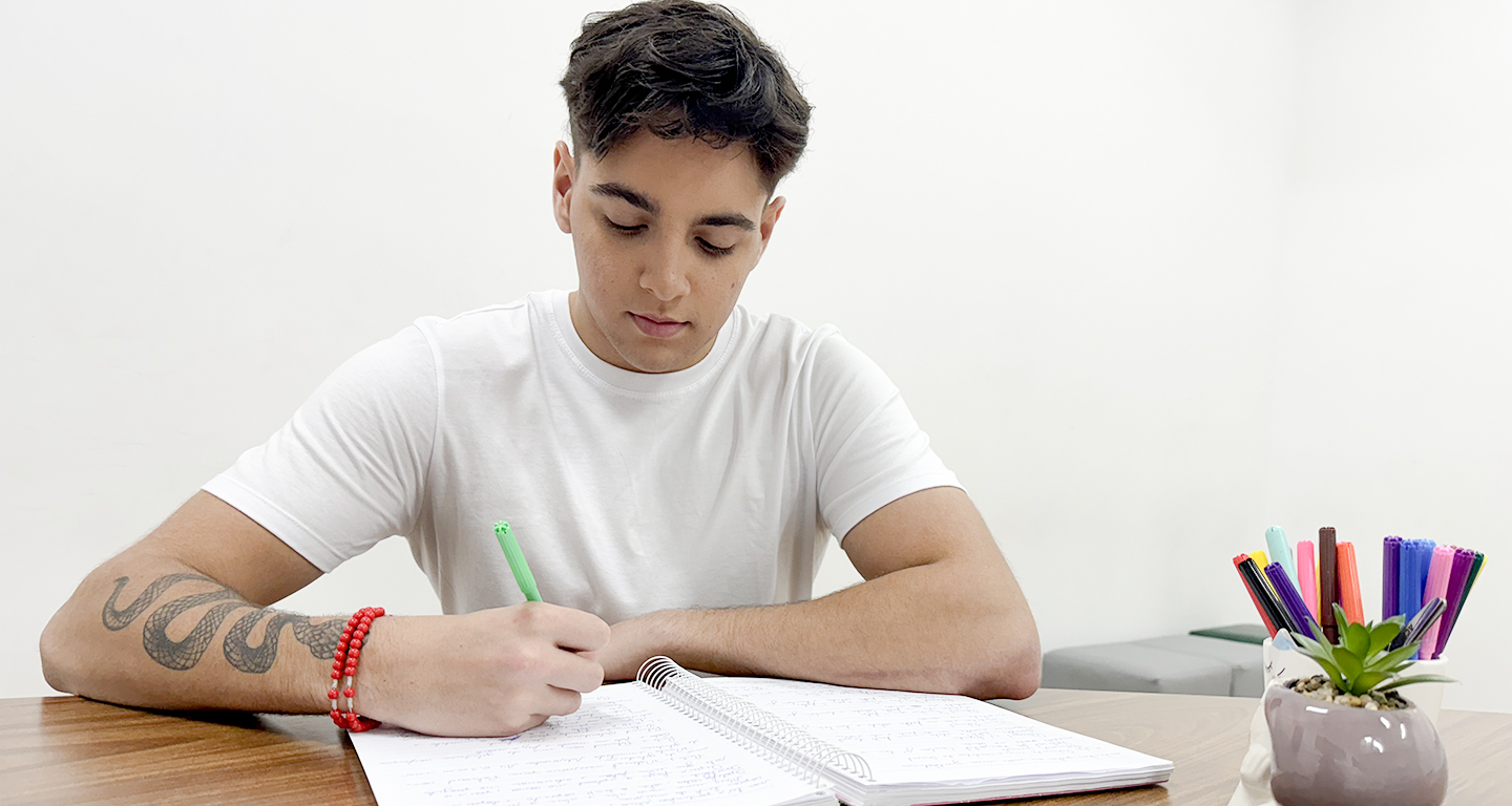 Jovem estudante sentado à mesa durante a preparação para o vestibular, escrevendo em um caderno com caneta verde. Ao lado, um suporte com canetas coloridas e uma pequena planta ajudam a compor um ambiente de estudo organizado e acolhedor, ideal para manter o foco e a disciplina.