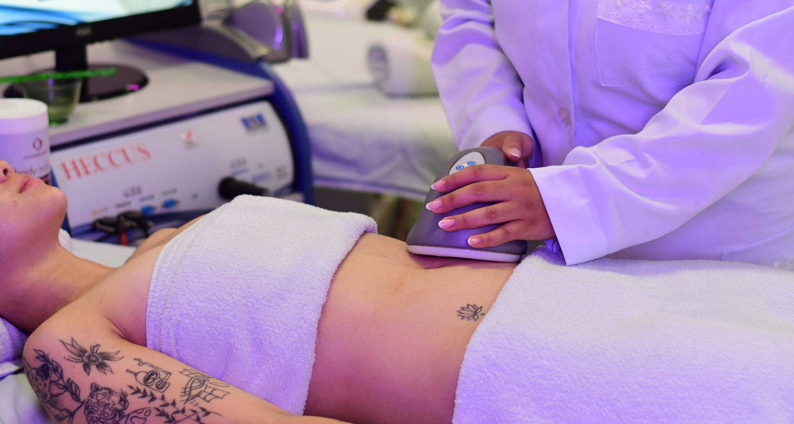 estudante de estética da Unoeste vestida com jaleco branco realiza procedimento estético com aparelho na região abdominal de uma paciente deitada, coberta com toalhas brancas, em uma clínica de estética.