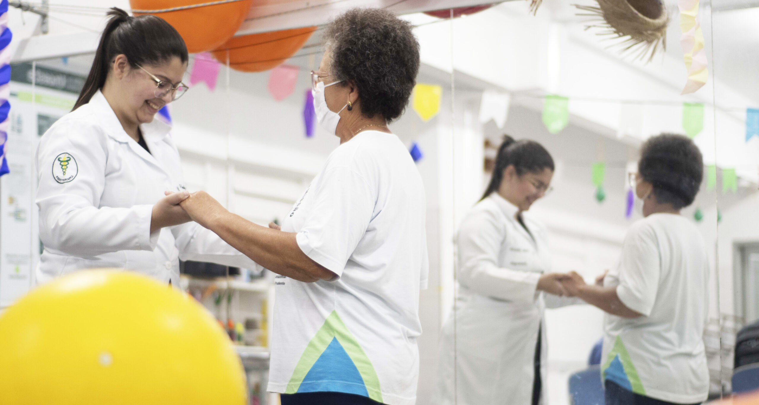 aluna de Fisioterapia da Unoeste vestida com jaleco branco sorri enquanto auxilia uma paciente idosa durante exercício em uma clínica de Fisioterapia. O ambiente é claro e está decorado com bandeirinhas coloridas, refletidas no espelho.