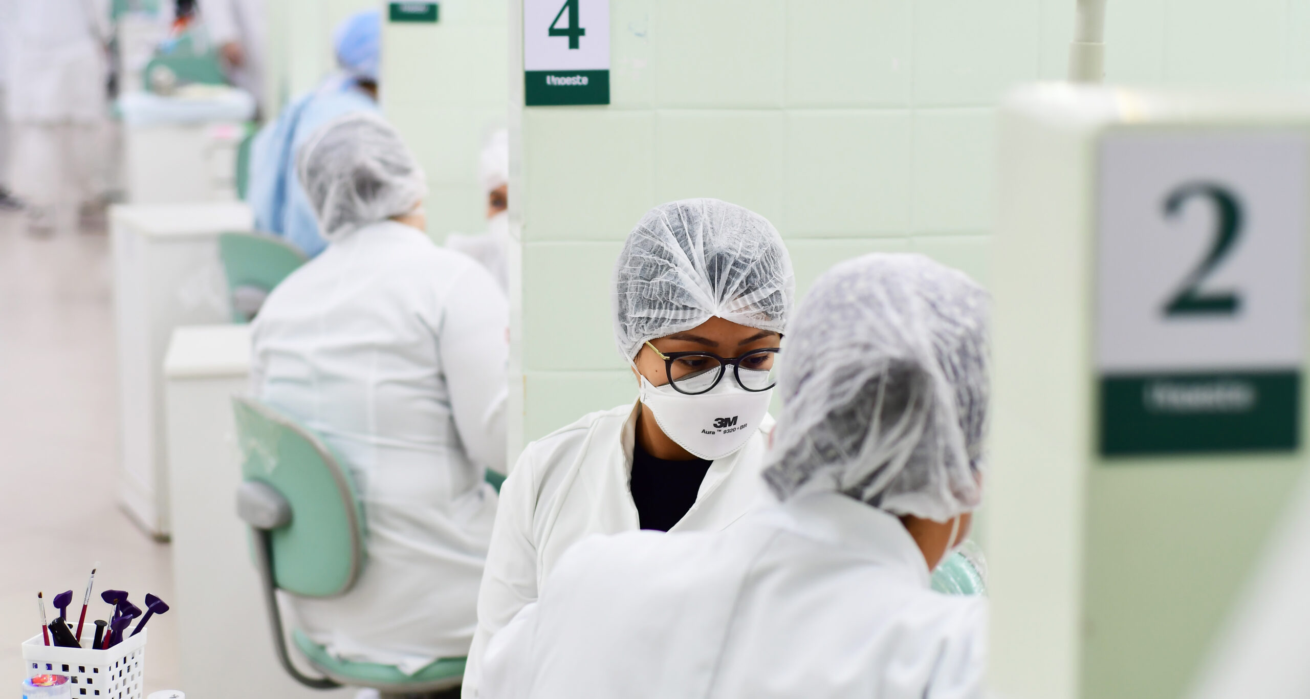 Ambiente clínico odontológico com várias cabines numeradas. Estudantes de Odontologia da Unoeste usam jalecos brancos, toucas descartáveis e máscaras faciais. Em destaque, duas alunas realizam atendimento odontológico enquanto se concentram no procedimento. Equipamentos e instrumentos dentários estão organizados ao fundo, em um ambiente limpo e bem iluminado.