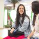 Ao fundo, área de convivência em faculdade particular. Em primeiro plano, duas estudantes usando mochila e bolsa sorriem enquanto conversam. 
