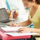 Uma jovem estudante concentra-se em escrever em um caderno, com um laptop aberto ao lado. A imagem transmite a dedicação de quem está se preparando ou preenchendo a inscrição do ProUni, buscando sua bolsa de estudos para o ensino superior e construindo um futuro de oportunidades.