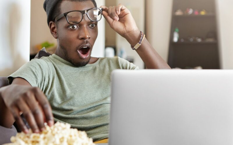 Na foto, um jovem negro pegando pipoca com uma mão e segurando os óculos de grau com a outra, boquiaberto e com a expressão de impressionando assistindo um filme de psicologia no notebook.