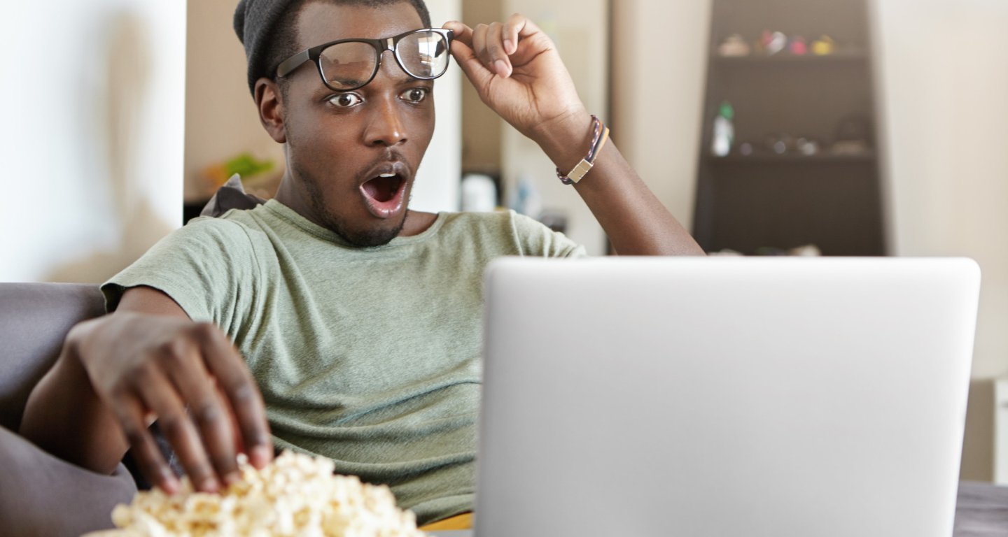 Na foto, um jovem negro pegando pipoca com uma mão e segurando os óculos de grau com a outra, boquiaberto e com a expressão de impressionando assistindo um filme de psicologia no notebook.