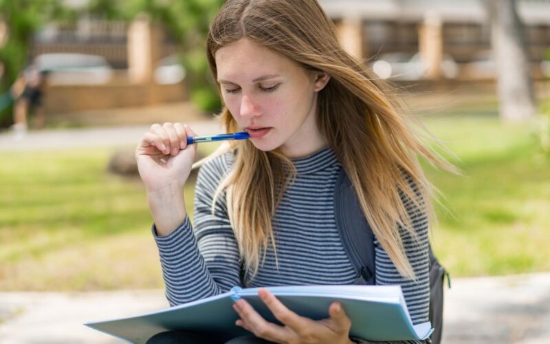 Ao fundo da imagem, uma universidade em desfoque. Enquanto a frente, no foco principal, uma jovem loira, segurando uma caneta próxima da boca e um caderno sobre as pernas avaliando a possibilidade de participar do vestibular como treineiro.