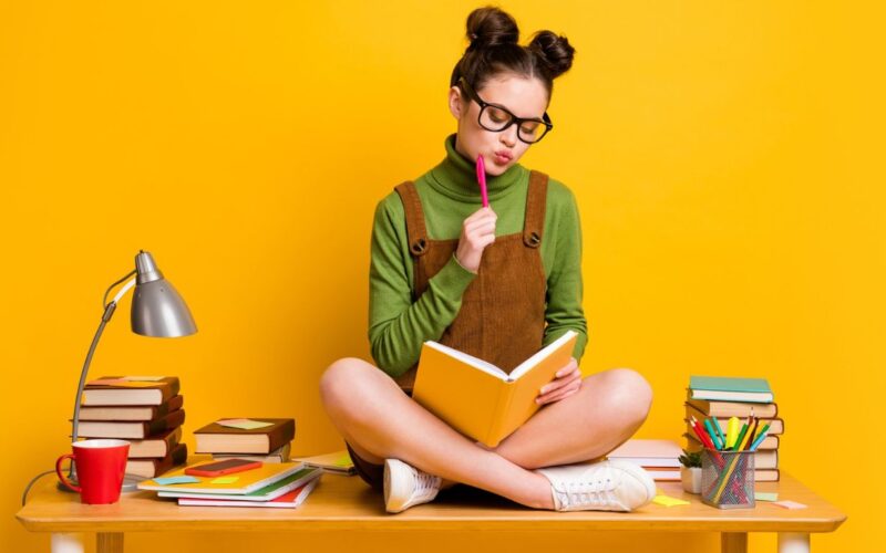 Na foto, uma jovem branca, sentada em cima de mesa, com as pernas cruzadas e segurando um caderno fazendo o método Cornell.