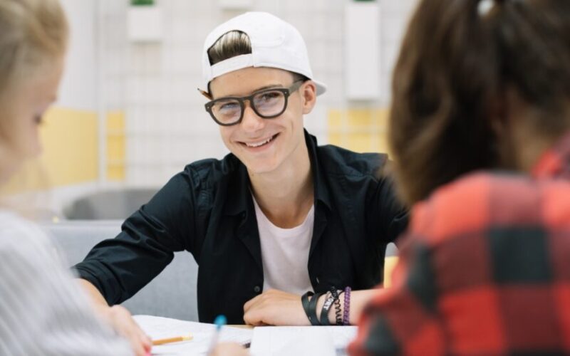 Aluno sorrindo enquanto está sentado em mesa de estudos, segurando caneta e fazendo resumo em caderno.