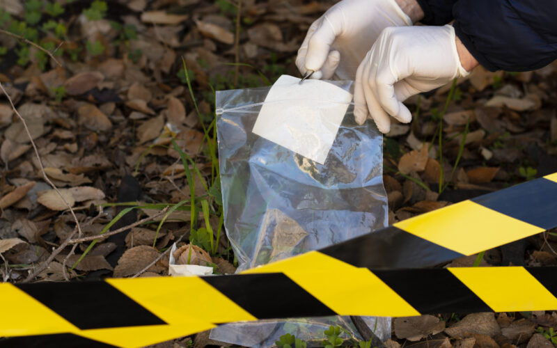 Perito criminal recolhendo uma evidência em uma cena de crime. Ele usa luvas, saco plástico e uma pinça e está em um local aberto com faixa sinalizando a ocorrência.