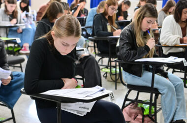 Sala de aula com vários jovens estudantes sentados em carteiras individuais, concentrados na leitura e resolução de uma prova escrita, com folhas de avaliação e canetas sobre as mesas durante a realização de um processo seletivo.