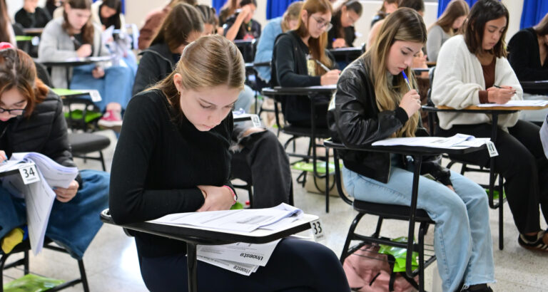 Sala de aula com vários jovens estudantes sentados em carteiras individuais, concentrados na leitura e resolução de uma prova escrita, com folhas de avaliação e canetas sobre as mesas durante a realização de um processo seletivo.