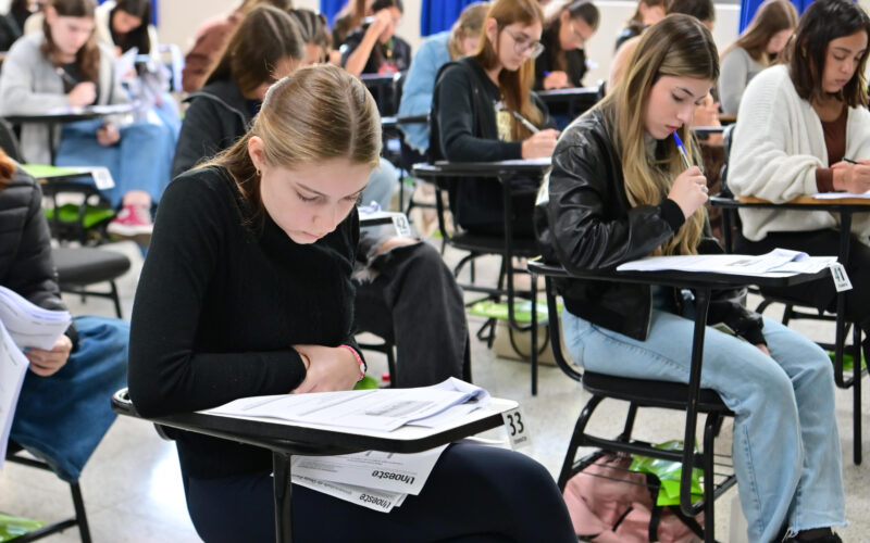 Sala de aula com vários jovens estudantes sentados em carteiras individuais, concentrados na leitura e resolução de uma prova escrita, com folhas de avaliação e canetas sobre as mesas durante a realização de um processo seletivo.