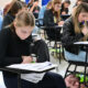 Sala de aula com vários jovens estudantes sentados em carteiras individuais, concentrados na leitura e resolução de uma prova escrita, com folhas de avaliação e canetas sobre as mesas durante a realização de um processo seletivo.