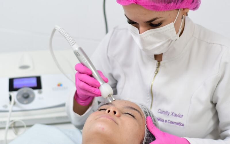 aluna do curso de Estética e Cosmética da Unoeste, com touca, luvas cor-de-rosa e jaleco branco, realizando procedimento no rosto de paciente. Há uma máquina de procedimento estético no fundo da imagem.