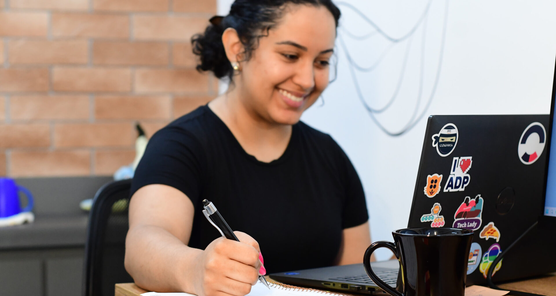Jovem estudante sorrindo em frente a um notebook enquanto faz anotações em um caderno.