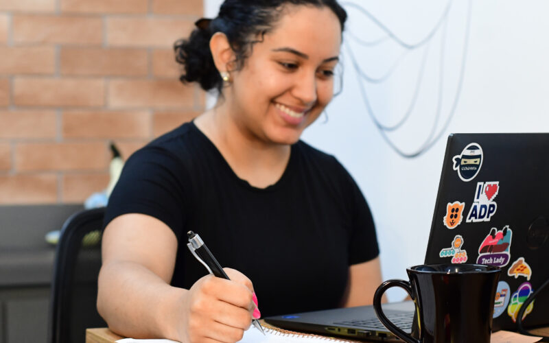 Jovem estudante sorrindo em frente a um notebook enquanto faz anotações em um caderno.