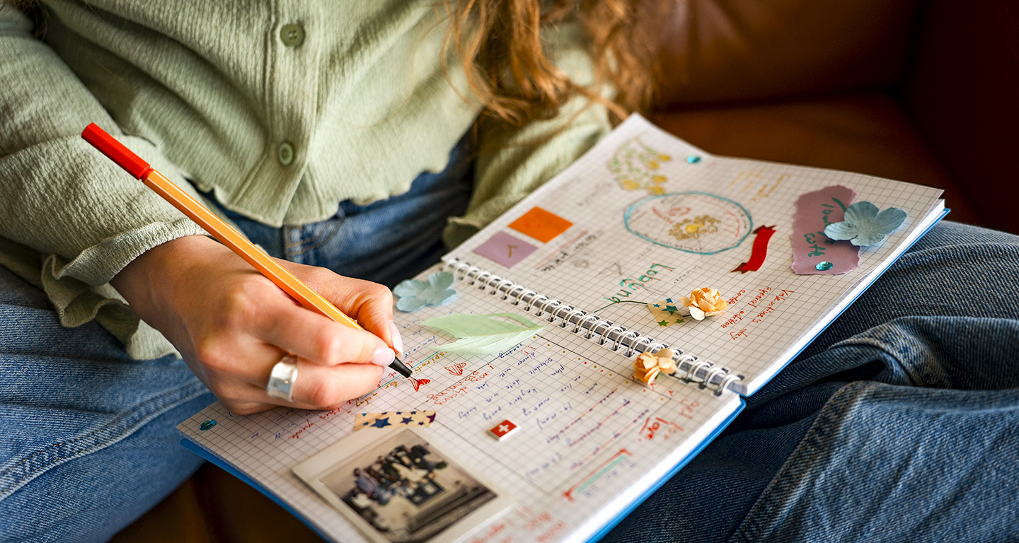 Uma pessoa sentada em um sofá, vestindo uma camisa verde-clara e calça jeans, escreve em um planner aberto. O planner é decorado com adesivos, recortes e uma foto. O foco está na mão da pessoa escrevendo com uma caneta laranja. Este é um exemplo visual de como um planner pode ser personalizado e usado para organização.