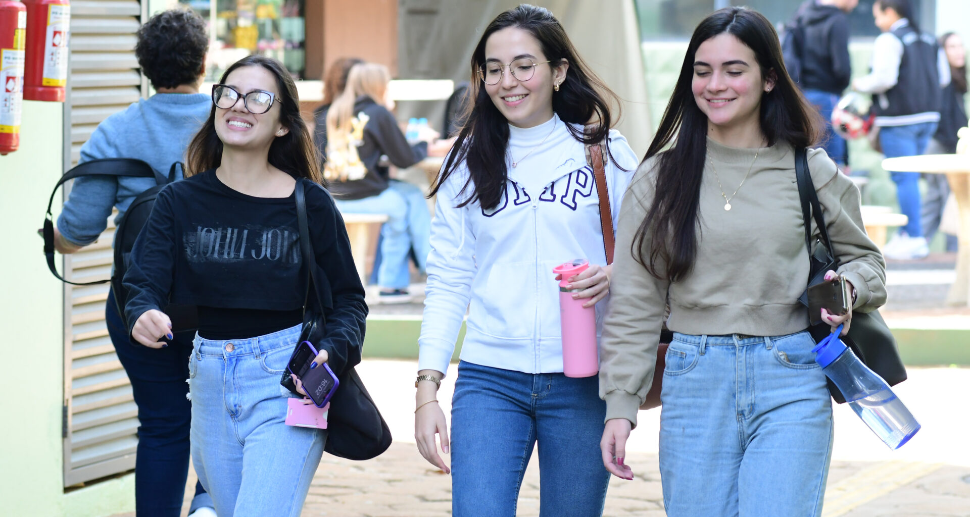 Três jovens estudantes da Unoeste sorridentes caminhando lado a lado no campus, carregando garrafas de água e mochilas, em um dia ensolarado. A cena reflete descontração e companheirismo no ambiente acadêmico.