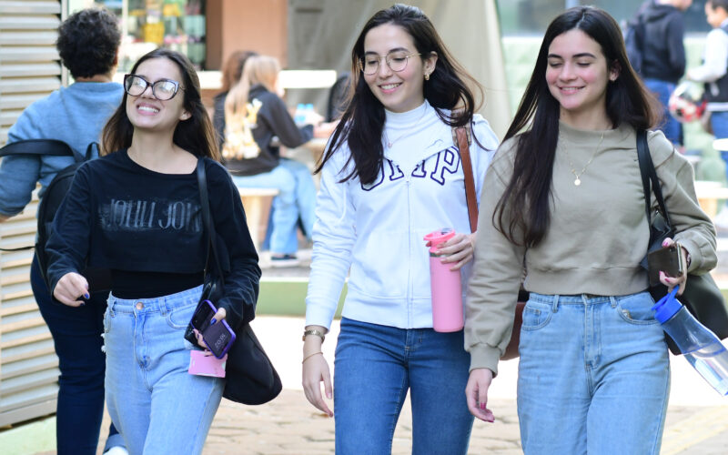 Três jovens estudantes da Unoeste sorridentes caminhando lado a lado no campus, carregando garrafas de água e mochilas, em um dia ensolarado. A cena reflete descontração e companheirismo no ambiente acadêmico.