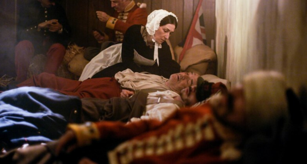 Cena do filme 'Florence Nightingale', onde a protagonista está ajudando um paciente debilitado, demonstrando compaixão e dedicação.