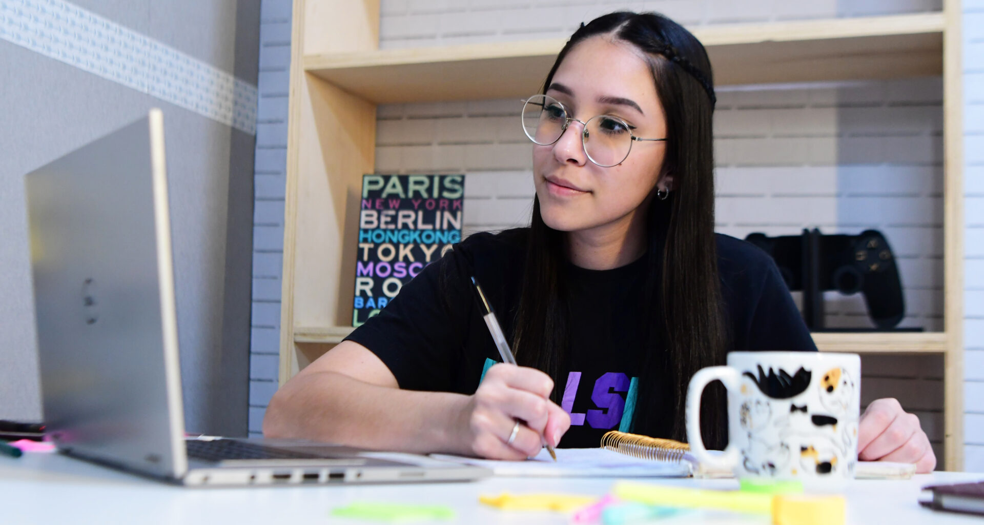 jovem garota estudando em um quarto com o auxílio de um notebook. Ela está sentada e em sua mesa contém vários objetos como canetas, bloquinhos de anotações, caderno e uma caneca.