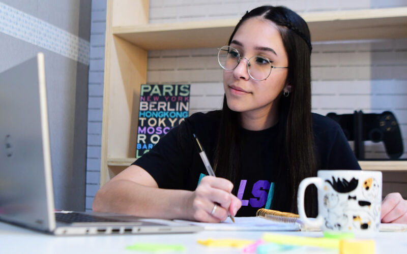 jovem garota estudando em um quarto com o auxílio de um notebook. Ela está sentada e em sua mesa contém vários objetos como canetas, bloquinhos de anotações, caderno e uma caneca.