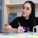 jovem garota estudando em um quarto com o auxílio de um notebook. Ela está sentada e em sua mesa contém vários objetos como canetas, bloquinhos de anotações, caderno e uma caneca.