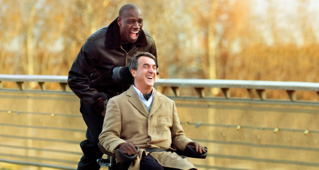 Imagem do filme 'Intocáveis', mostrando o personagem Philippe em uma cadeira de rodas, rindo ao lado de Driss, seu cuidador. 
