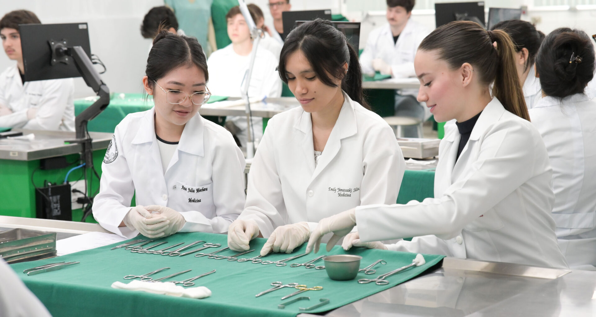 Estudantes de Medicina em laboratório participam de atividade prática, organizando e manuseando instrumentos cirúrgicos sobre bancada, utilizando jalecos e luvas, em ambiente acadêmico com outros alunos ao fundo.