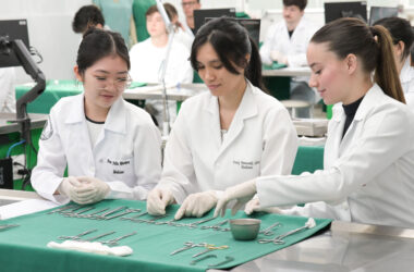 Estudantes de Medicina em laboratório participam de atividade prática, organizando e manuseando instrumentos cirúrgicos sobre bancada, utilizando jalecos e luvas, em ambiente acadêmico com outros alunos ao fundo.