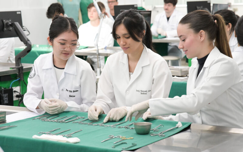 Estudantes de Medicina em laboratório participam de atividade prática, organizando e manuseando instrumentos cirúrgicos sobre bancada, utilizando jalecos e luvas, em ambiente acadêmico com outros alunos ao fundo.