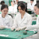 Estudantes de Medicina em laboratório participam de atividade prática, organizando e manuseando instrumentos cirúrgicos sobre bancada, utilizando jalecos e luvas, em ambiente acadêmico com outros alunos ao fundo.