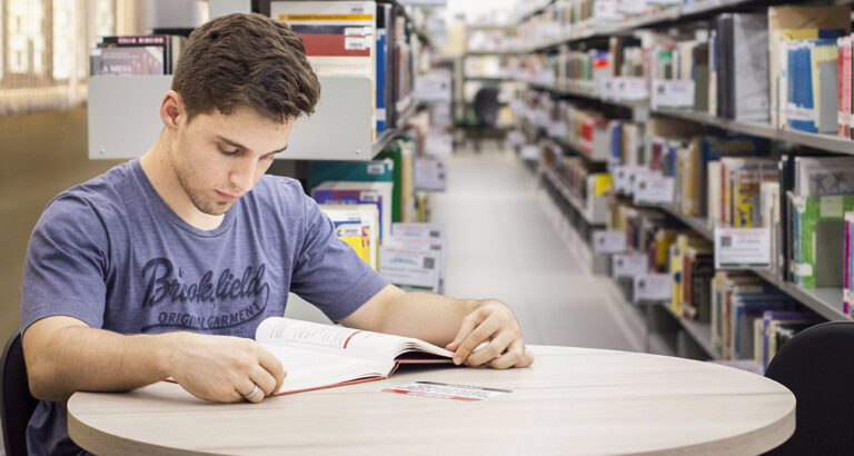 Homem jovem sentado em uma mesa redonda em uma biblioteca, lendo um livro. Ele está usando uma camiseta azul escura com letras brancas e tem cabelo castanho curto. A mesa é de cor clara e o livro está aberto à sua frente. No fundo, as prateleiras da biblioteca estão cheias de livros, criando um cenário acadêmico. A biblioteca está bem iluminada e o foco está no jovem e no livro, enquanto o fundo está ligeiramente desfocado. O jovem parece concentrado em um tipo de leitura reflexiva.