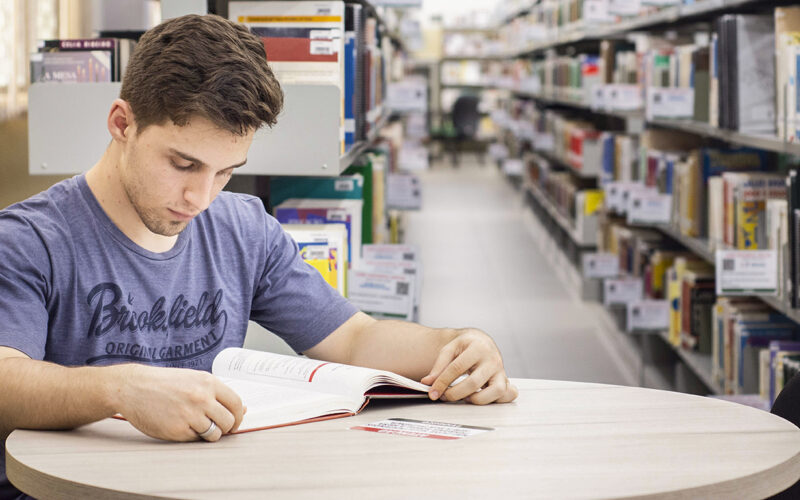 Homem jovem sentado em uma mesa redonda em uma biblioteca, lendo um livro. Ele está usando uma camiseta azul escura com letras brancas e tem cabelo castanho curto. A mesa é de cor clara e o livro está aberto à sua frente. No fundo, as prateleiras da biblioteca estão cheias de livros, criando um cenário acadêmico. A biblioteca está bem iluminada e o foco está no jovem e no livro, enquanto o fundo está ligeiramente desfocado. O jovem parece concentrado em um tipo de leitura reflexiva.