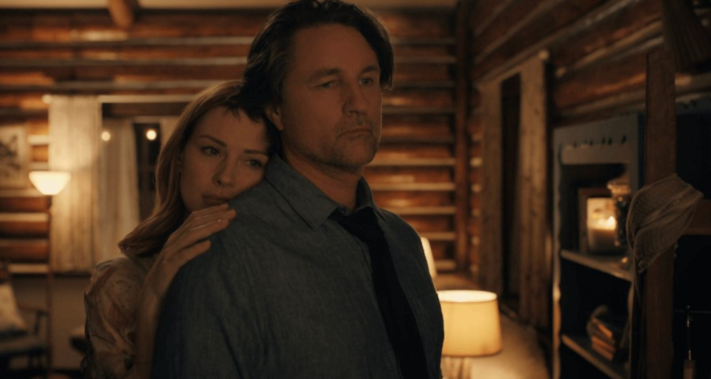 Captura de cena da série Virgin River, na qual a protagonista Melinda Monroe apoia seu rosto nas costas de Jack Sheridan, que olha para frente. Ao fundo, uma sala em casa de madeira.