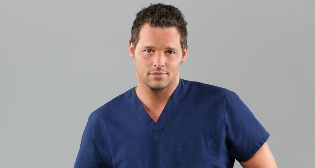 Um retrato de Justin Chambers como Alex Karev em Grey's Anatomy. Ele está usando um uniforme azul marinho e olhando para a câmera com uma expressão neutra. O fundo é cinza sólido.