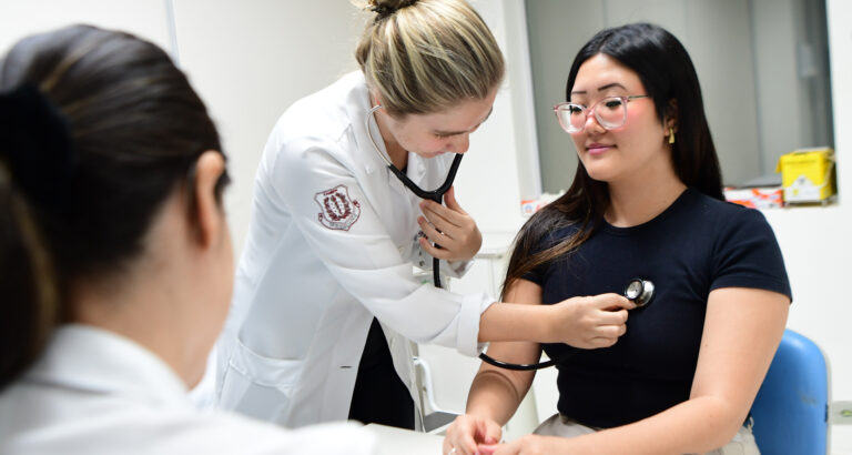 Estudantes de Medicina da Unoeste realizando prática clínica em um ambiente hospitalar, onde uma aluna utiliza um estetoscópio para examinar uma paciente, ilustrando o aprendizado prático do curso.