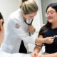 Estudantes de Medicina da Unoeste realizando prática clínica em um ambiente hospitalar, onde uma aluna utiliza um estetoscópio para examinar uma paciente, ilustrando o aprendizado prático do curso.