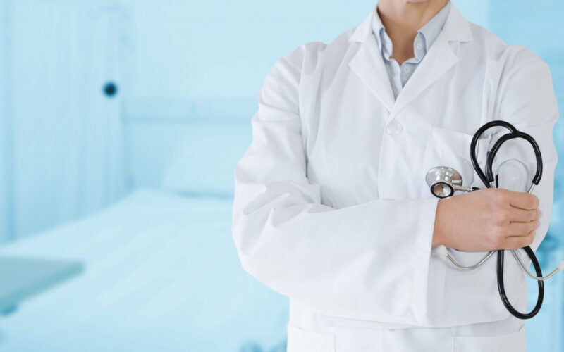 Médico de jaleco branco segurando estetoscópio em ambiente hospitalar.