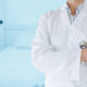 Médico de jaleco branco segurando estetoscópio em ambiente hospitalar.