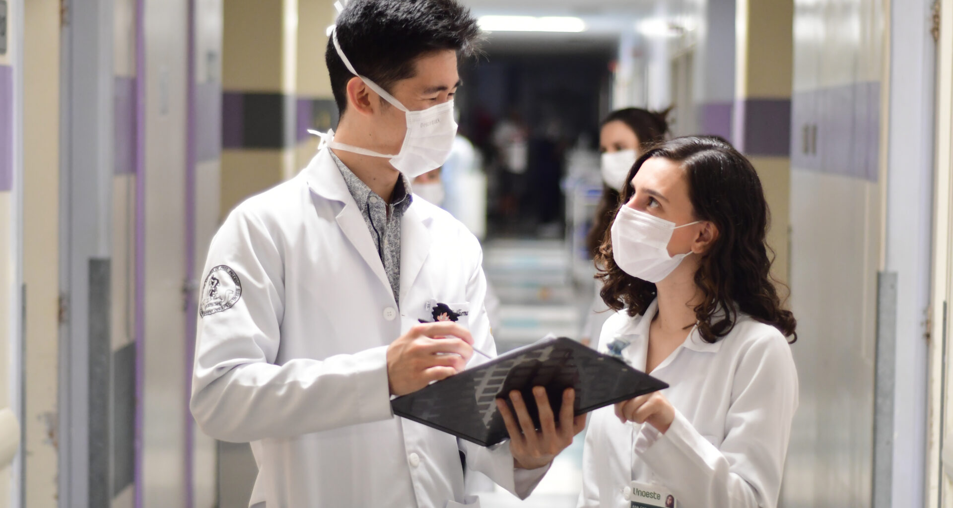 Dois alunos de Medicina da Unoeste, usando jalecos brancos e máscaras de proteção, conversam em um corredor de hospital. Um deles segura um prontuário, enquanto discutem informações médicas de forma colaborativa. Ao fundo, é possível ver o ambiente hospitalar, com iluminação clara e movimentação de pessoas.