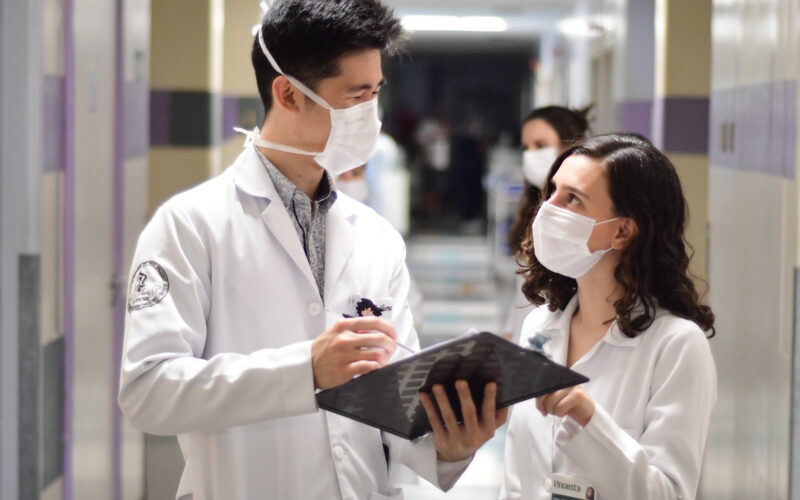 Dois alunos de Medicina da Unoeste, usando jalecos brancos e máscaras de proteção, conversam em um corredor de hospital. Um deles segura um prontuário, enquanto discutem informações médicas de forma colaborativa. Ao fundo, é possível ver o ambiente hospitalar, com iluminação clara e movimentação de pessoas.