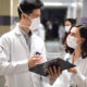 Dois alunos de Medicina da Unoeste, usando jalecos brancos e máscaras de proteção, conversam em um corredor de hospital. Um deles segura um prontuário, enquanto discutem informações médicas de forma colaborativa. Ao fundo, é possível ver o ambiente hospitalar, com iluminação clara e movimentação de pessoas.