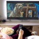 Mulher sentada em uma poltrona aconchegante, segurando um controle remoto e uma tigela de pipoca, assistindo a um filme de cachorro em uma TV de tela grande. A cena mostra um cachorro em destaque na tela, enquanto o ambiente ao redor é decorado com plantas e móveis de madeira, criando uma atmosfera acolhedora e relaxante.