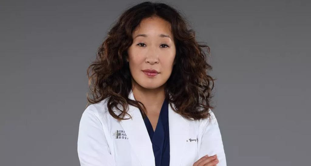 Um retrato de Sandra Oh como Cristina Yang em Grey's Anatomy. Ela está usando um jaleco branco sobre um uniforme azul marinho e tem os braços cruzados. Seu cabelo castanho está solto e ela está olhando para a câmera com uma expressão neutra. O fundo é cinza sólido.