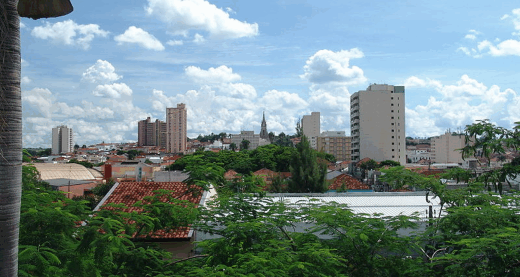 Vista aérea do Guarujá, SP. Uma longa estrada reta corta a paisagem, ladeada por floresta densa à esquerda e edifícios comerciais à direita. Ao fundo, a cidade se estende com prédios brancos e o oceano azul. Montanhas verdes completam a cena sob um céu claro.