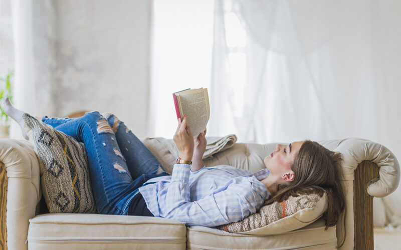 Mulher relaxando em um sofá enquanto lê um livro de romance com médico. O ambiente é iluminado e aconchegante, representando momentos de leitura e descanso.