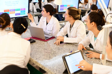 Estudantes de Medicina da Unoeste em um ambiente de aprendizado interativo, usando laptops e tablets enquanto discutem em grupo, com telas ao fundo exibindo conteúdos relacionados ao tratamento de pacientes com Her-2 positivo.